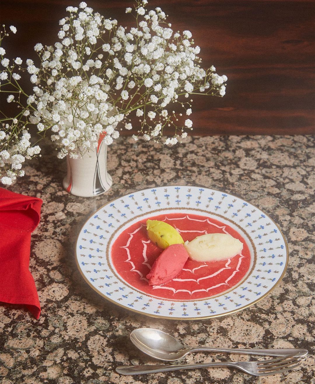 Overhead shot of a decorative plate featuring pink soup with dollops of yellow and cream sorbet, garnished with baby's breath flowers.