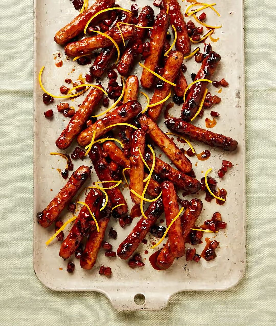 Overhead shot of glazed cocktail sausages arranged on a serving tray with candied orange peel and dark red sauce.