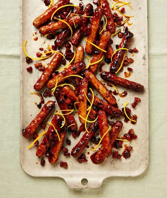 Overhead shot of glazed cocktail sausages arranged on a serving tray with candied orange peel and dark red sauce.