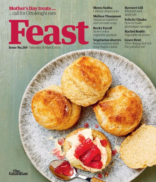 Close-up of a magazine cover featuring a pile of scones, some topped with cream and red jam, set on a gray speckled plate.