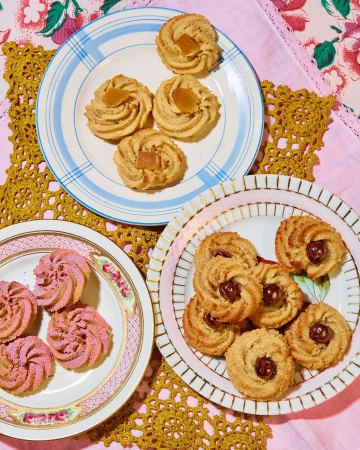 Three plates of homemade cookies with various toppings sit on a crocheted placemat and a pink floral cloth, photographed in natural light.