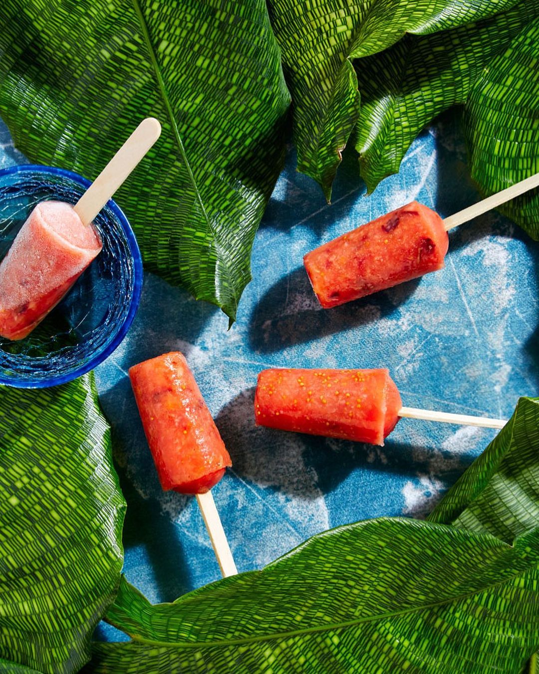 Overhead shot of four strawberry popsicles on sticks, one in blue bowl, surrounded by green patterned leaves on blue surface.