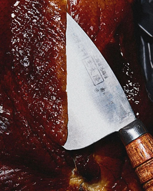 Close-up shot of a knife cutting into glistening, reddish-brown roast meat. Wooden handle of knife, stainless steel blade with Japanese text visible.