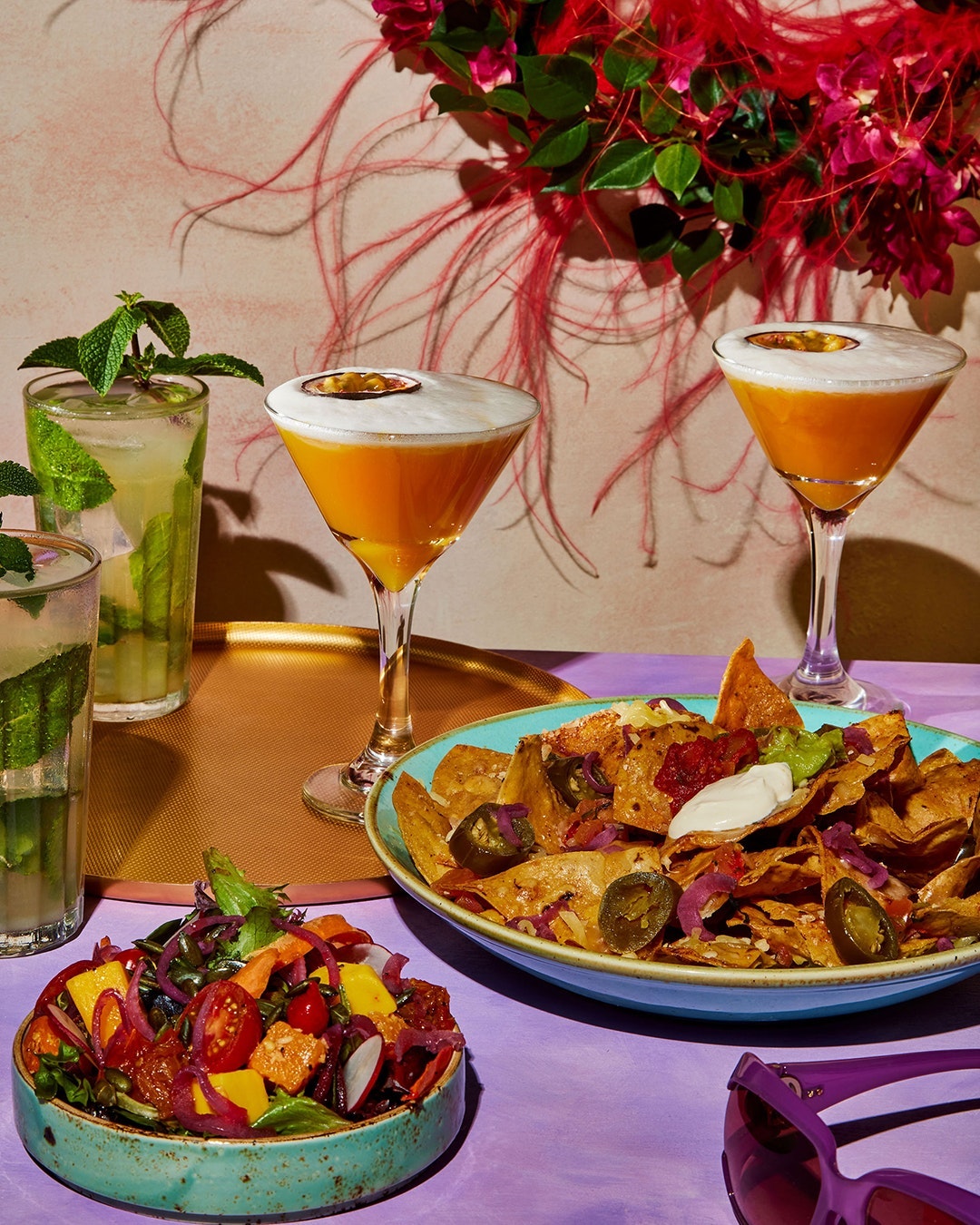 Cocktails and appetizers on a table, featuring drinks with mint and orange cocktails with a white foamy top, and a plate of nachos.