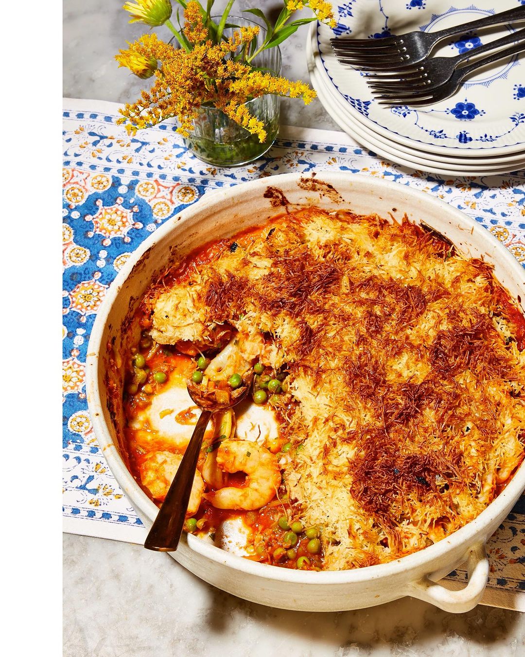Overhead shot of a baked shrimp casserole dish with a golden crust, garnished with peas, with flowers, plates, and flatware in background.