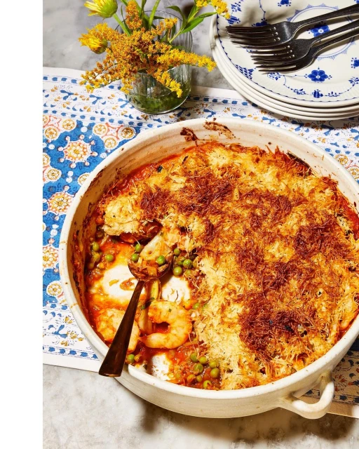 Overhead shot of a baked shrimp casserole dish with a golden crust, garnished with peas, with flowers, plates, and flatware in background.