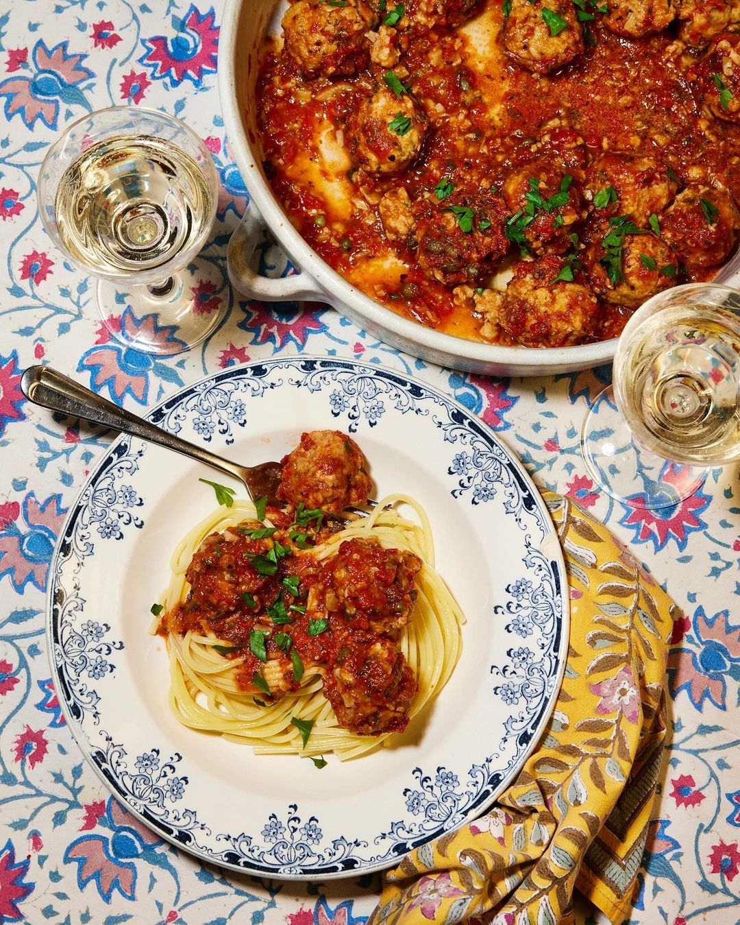 Overhead shot of spaghetti with meatballs in tomato sauce on a floral tablecloth with a casserole dish and drinks.