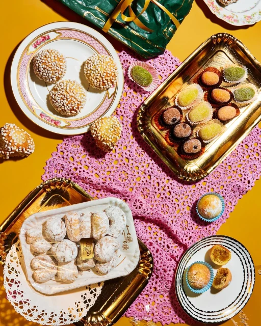 Overhead shot of an assortment of sweets and pastries on plates and trays, arranged on a yellow surface with a pink doily.