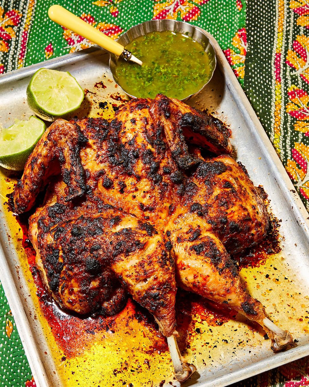 Overhead shot of a spiced, butterflied roast chicken on a baking sheet with lime wedges and dipping sauce against a patterned cloth.