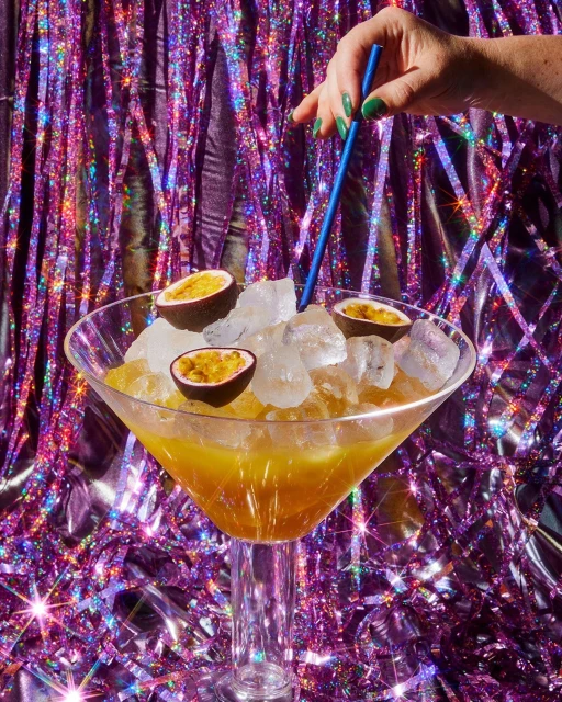 Cocktail in a large glass with ice, passion fruit, and a straw being held by a hand with green nail polish against a shiny backdrop.
