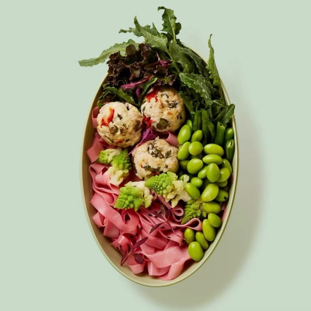 Overhead shot of a colorful salad bowl with pasta, leafy greens, meatballs, and edamame beans on a light green background.