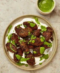 Overhead view of fried fritters drizzled with green herb sauce resting on yogurt inside a ceramic plate.