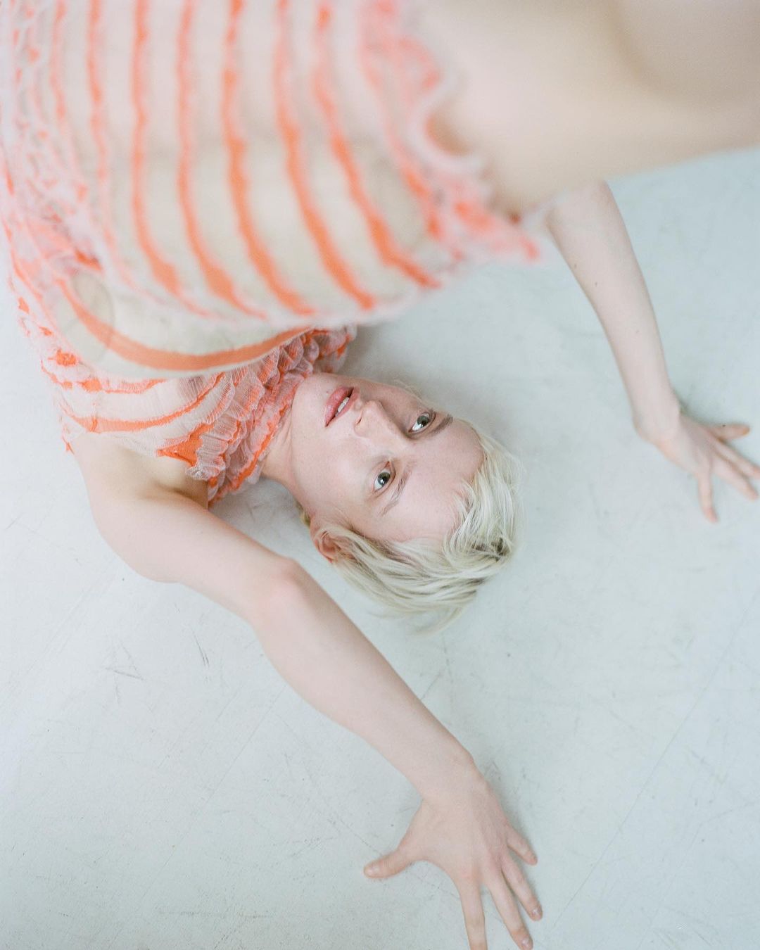 Woman with short blonde hair lies on her back in a light orange and white dress on a light grey surface in a studio setting.