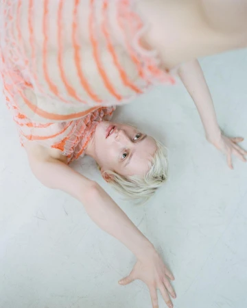 Woman with short blonde hair lies on her back in a light orange and white dress on a light grey surface in a studio setting.