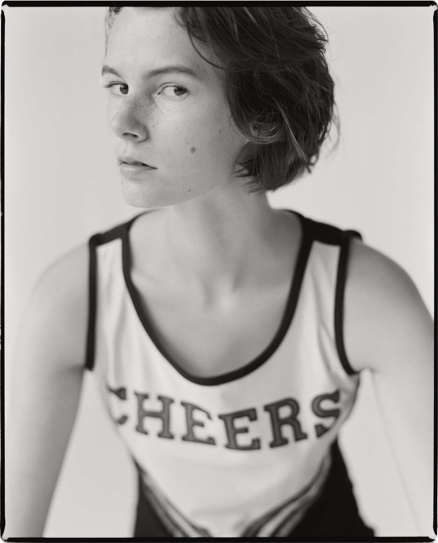 Monochrome portrait of a young woman with short hair wearing a cheerleading top, gazing toward the camera against a plain background.