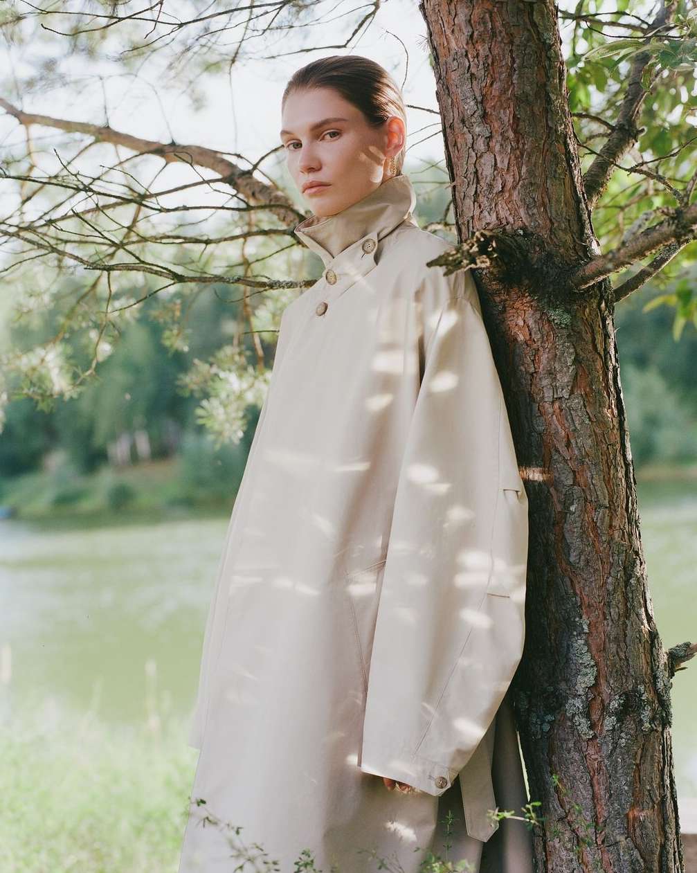 Woman in beige trench coat poses by tree against lake backdrop, natural light, neutral tones, serene, fashion editorial style.