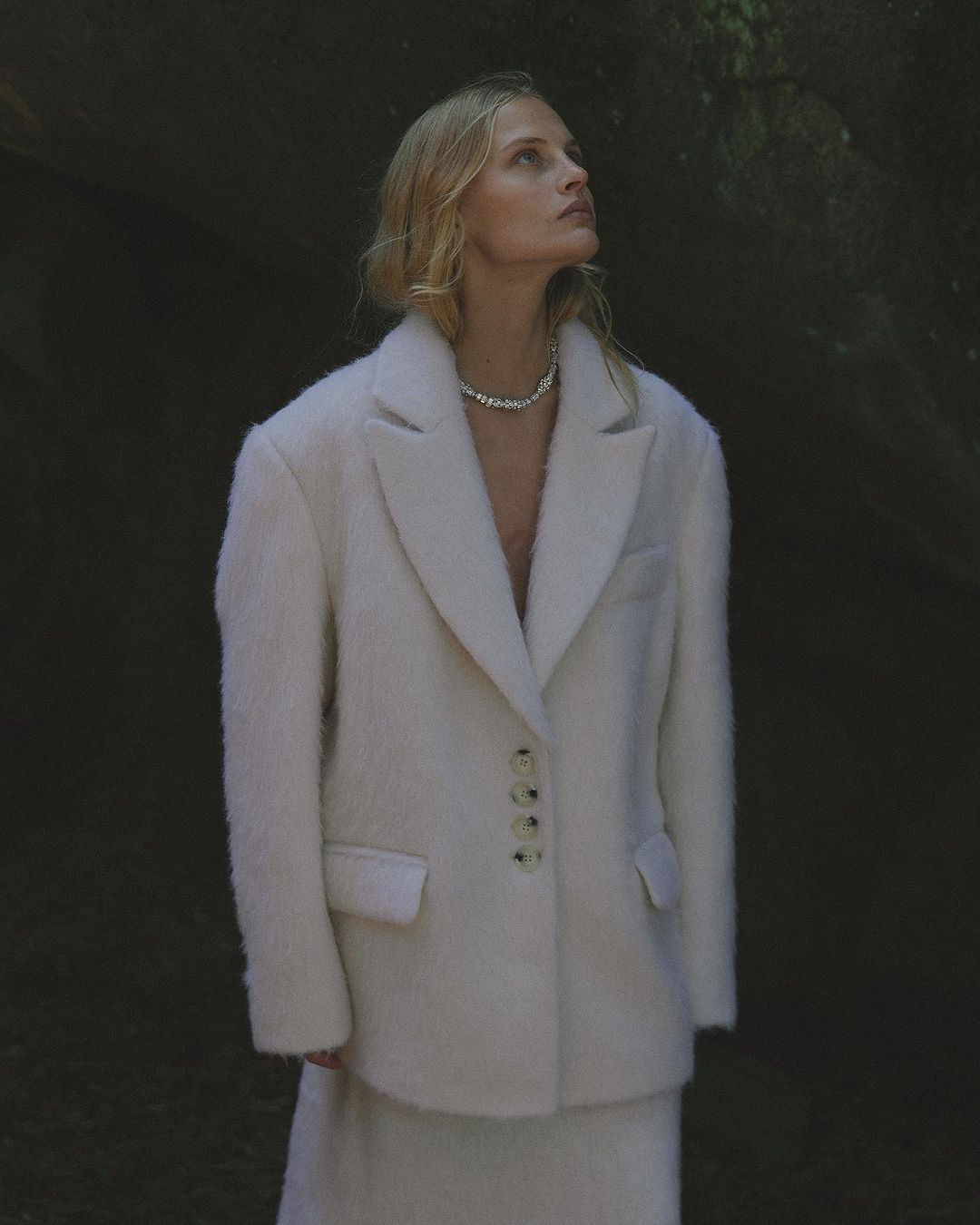 Model in a cream fuzzy textured blazer and necklace looking up outdoors by Luba Kozorezova