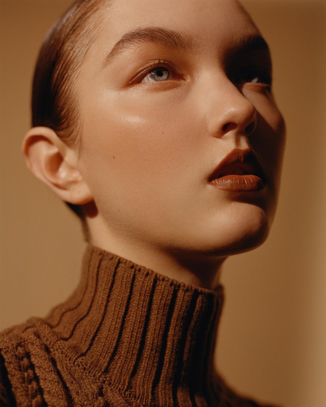 Close-up portrait of a young woman with blue eyes and brown lipstick wearing a brown knit turtleneck sweater, looking up.