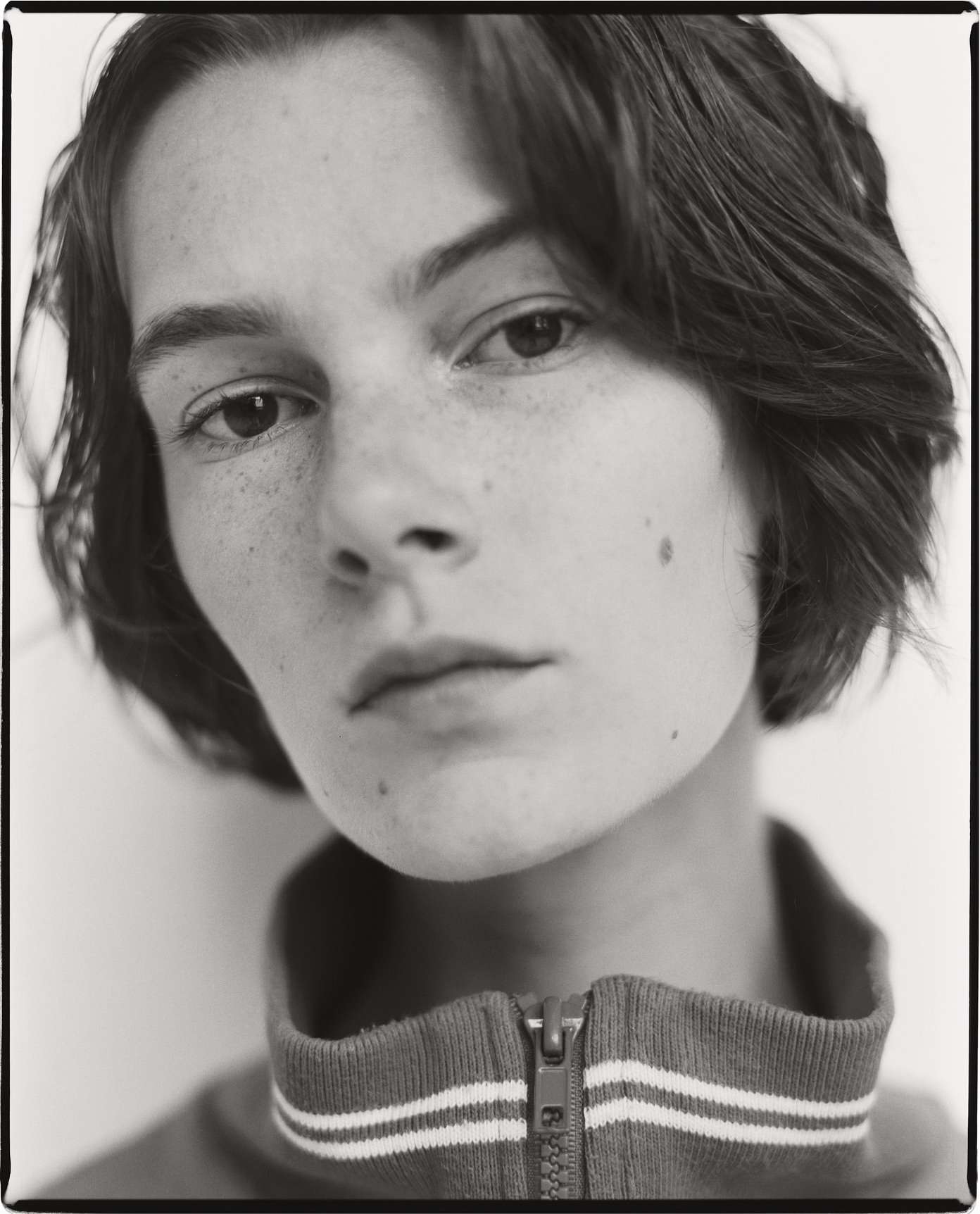 Monochrome portrait of a fair-skinned young adult woman with freckles and short brown hair wearing a zipped track jacket against white.