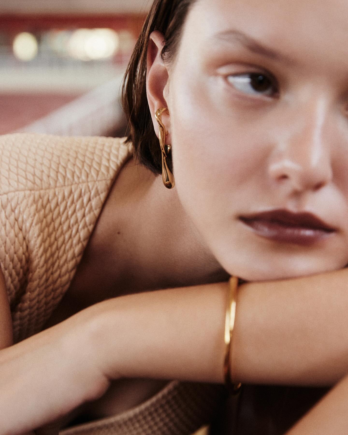 Close-up of a young adult female model with an elegant gold earring and matching bracelet, set against a blurred background.