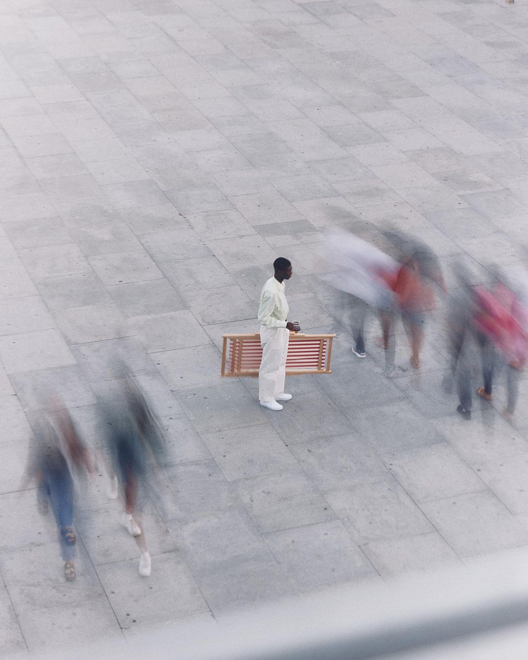 High-angle view of a black man in casual white attire standing alone near a bench as blurred people walk by on a grey-paved plaza.