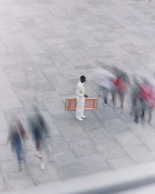 High-angle view of a black man in casual white attire standing alone near a bench as blurred people walk by on a grey-paved plaza.