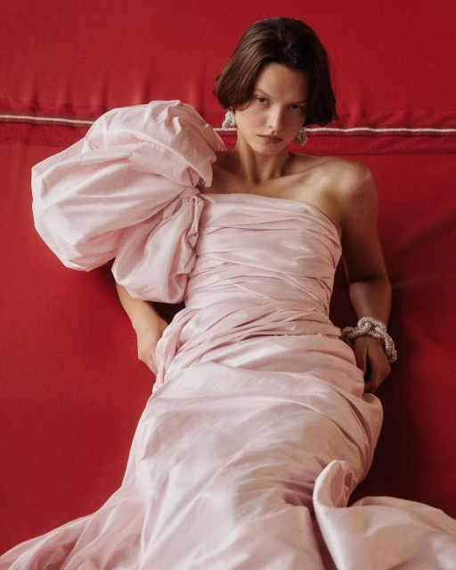 Young woman poses in a draped light pink one-shoulder gown against a red background, wearing ornate earrings and a bracelet.