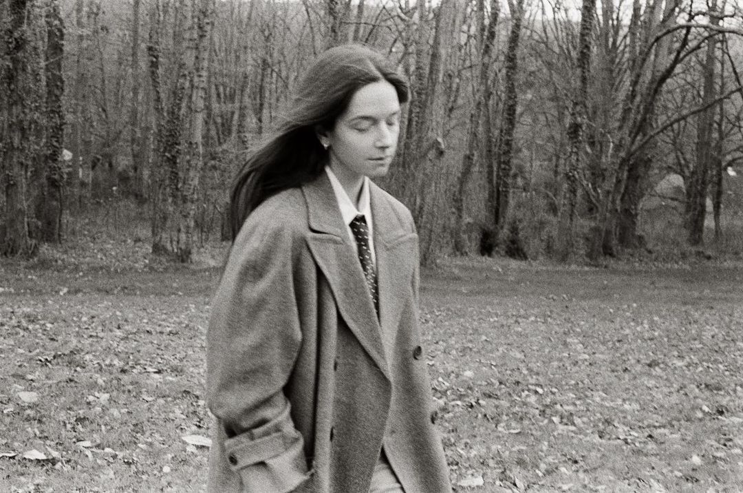 Monochrome portrait of a young woman wearing an overcoat and tie, standing outdoors in front of trees, with a serene expression.