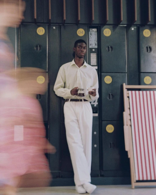 Stylish Black man in off-white outfit holds glasses, standing near lockers and striped folding chair in an indoor setting with soft light.