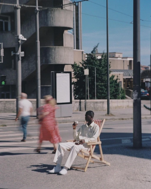 Young man in casual clothes relaxes in a beach chair on a city sidewalk, holding a cup, with blurred pedestrians and buildings behind.
