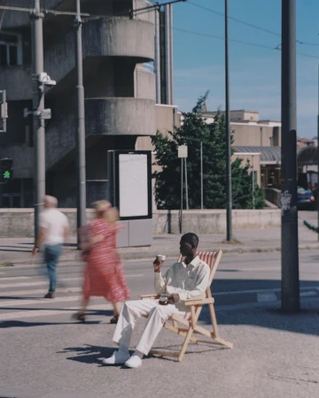 Young man in casual clothes relaxes in a beach chair on a city sidewalk, holding a cup, with blurred pedestrians and buildings behind.