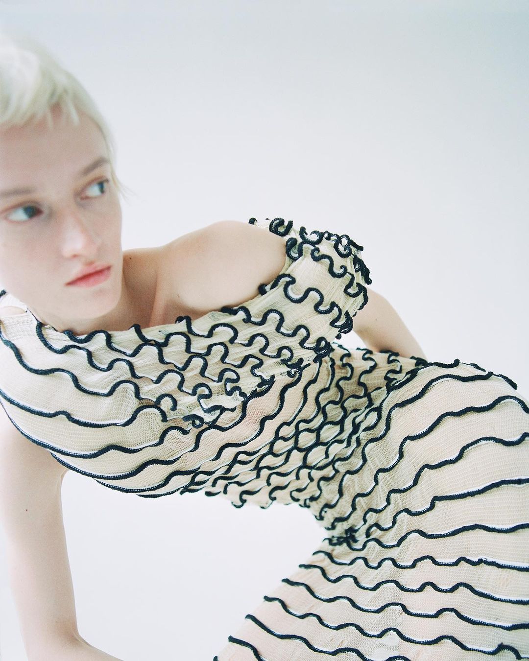 Blonde woman in a white dress with black ruffles posing against a white background in a fashion setting, looking left.