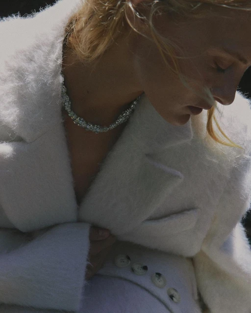 Close-up of a blonde adult female wearing a white coat and an elaborate jewel-encrusted necklace, set against a dark, shadowed background.