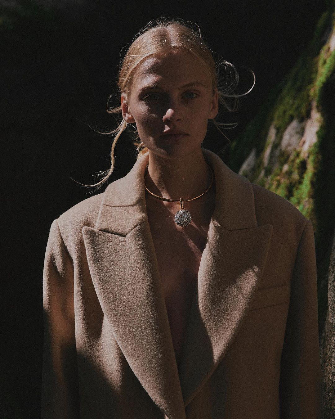 Woman with blonde hair wearing a beige coat and a gold necklace with a pendant, posing outdoors in natural light against a mossy rock.