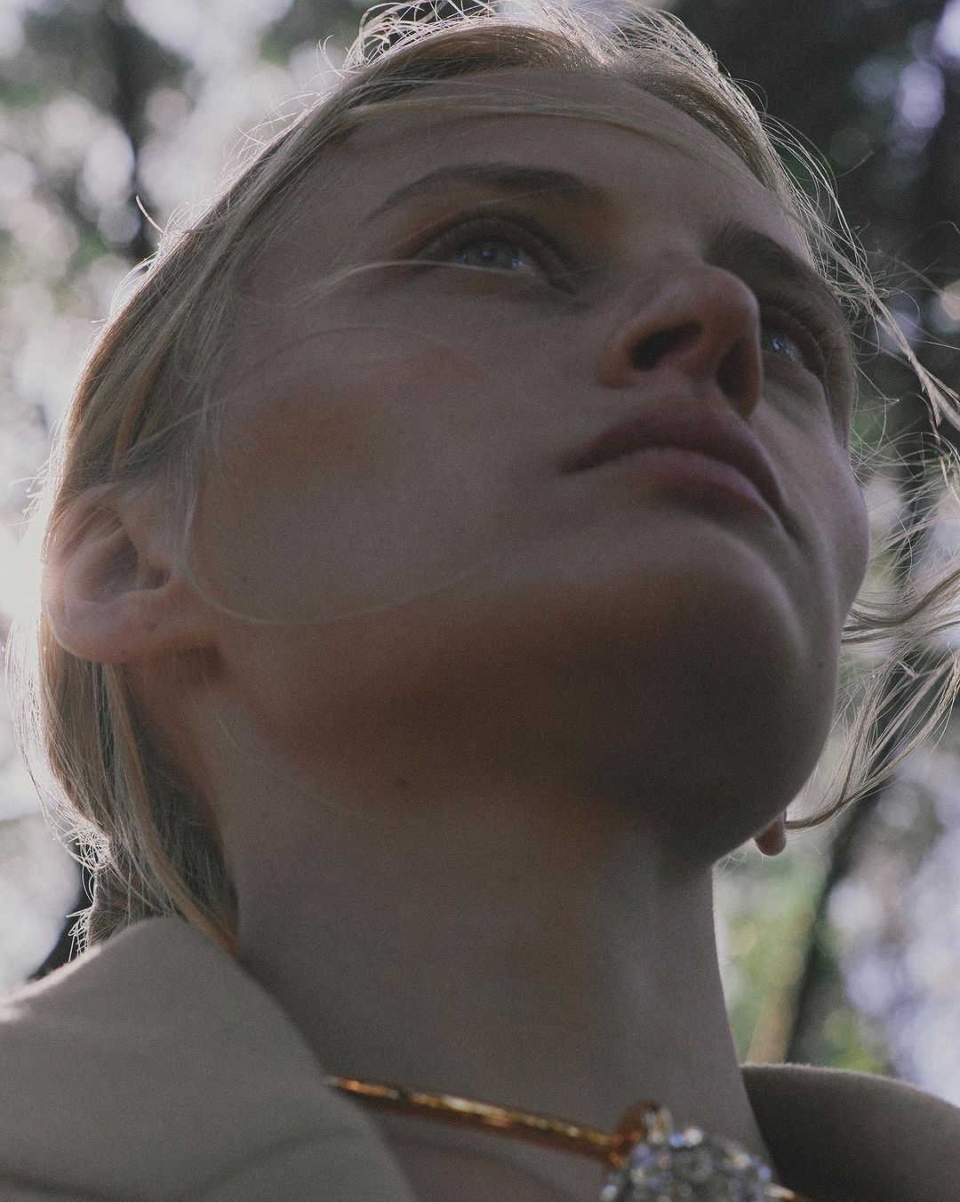 Close-up of a fair-skinned woman looking upwards with light blonde hair, wearing a gold necklace with jewel, against a blurred forest backdrop.