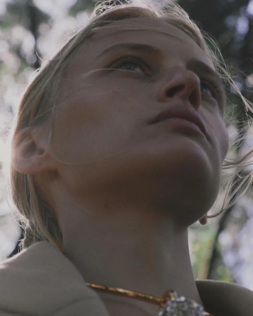 Close-up of a fair-skinned woman looking upwards with light blonde hair, wearing a gold necklace with jewel, against a blurred forest backdrop.