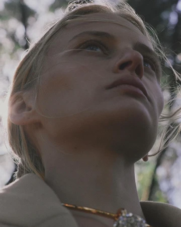 Close-up of a fair-skinned woman looking upwards with light blonde hair, wearing a gold necklace with jewel, against a blurred forest backdrop.