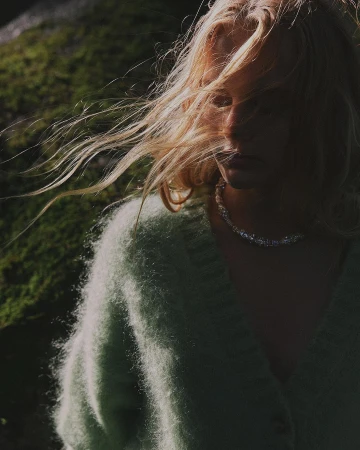 Portrait of a woman with windblown blonde hair wearing a fuzzy mint green cardigan and a beaded necklace against a dark mossy background.