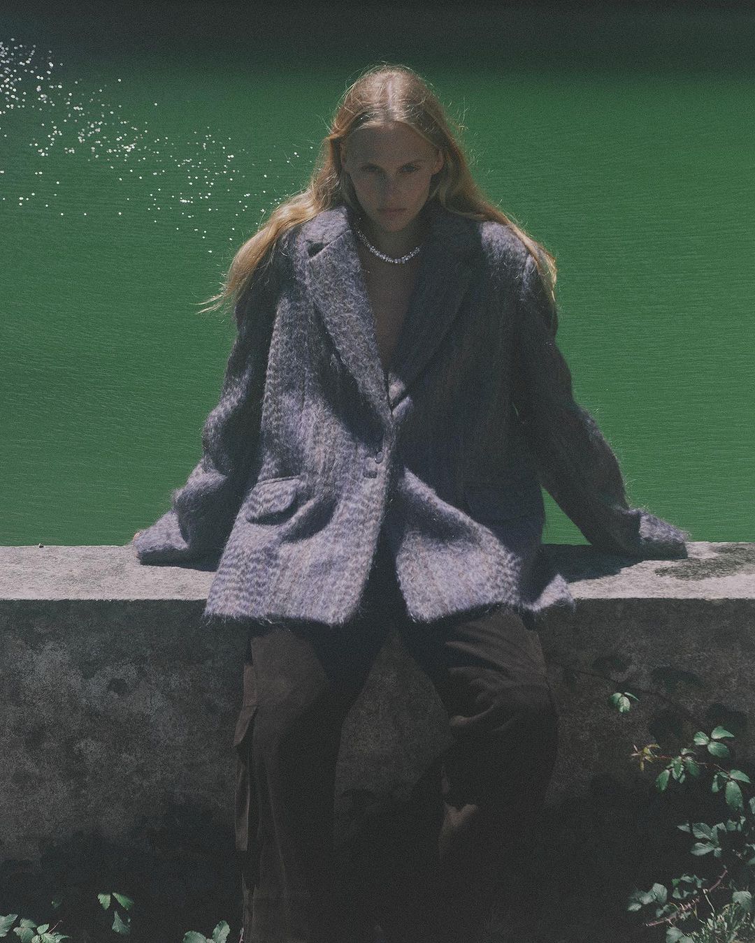A young woman with long blond hair sits on a ledge wearing a grey blazer and brown pants in front of a bright green pond outdoors.