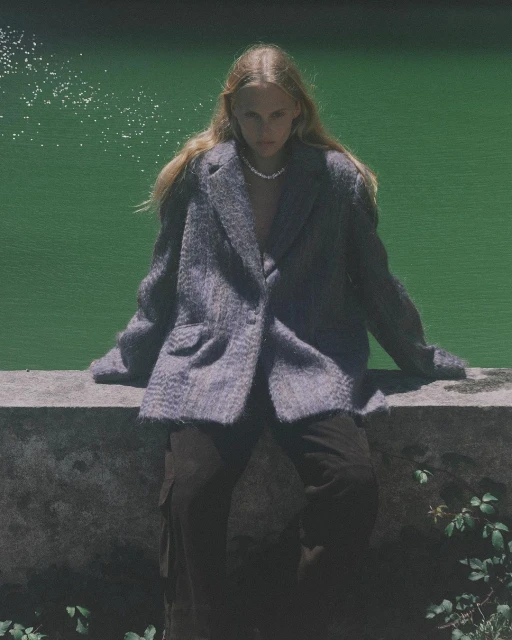 A young woman with long blond hair sits on a ledge wearing a grey blazer and brown pants in front of a bright green pond outdoors.