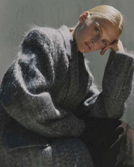 A young adult woman poses in a grey, textured coat. The portrait showcases a serene expression and soft, diffused lighting.