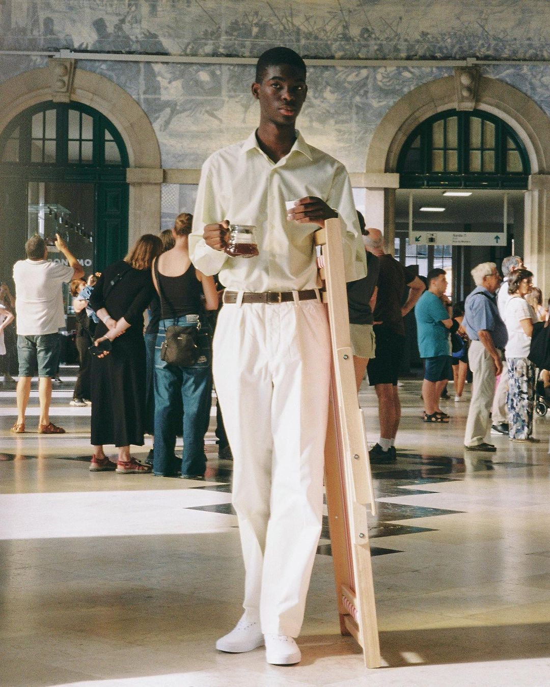Young Black man stands in train station with drink and folded photography ladder, wearing light green shirt and white pants, surrounded by travelers.