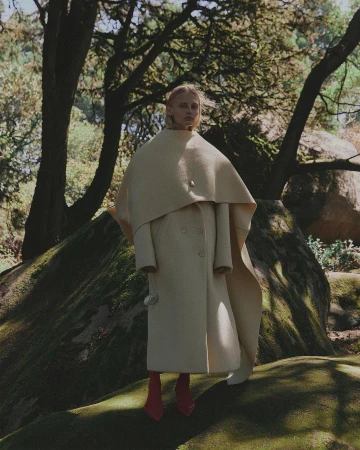 Fashion shot of a female model in an oversized cream coat and bright boots posing outside amongst green mossy rocks and trees.