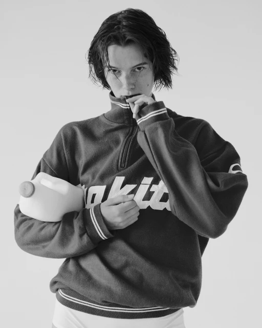 Monochrome portrait of a young adult holding a plastic milk jug wearing a branded quarter-zip sweatshirt against a plain background.