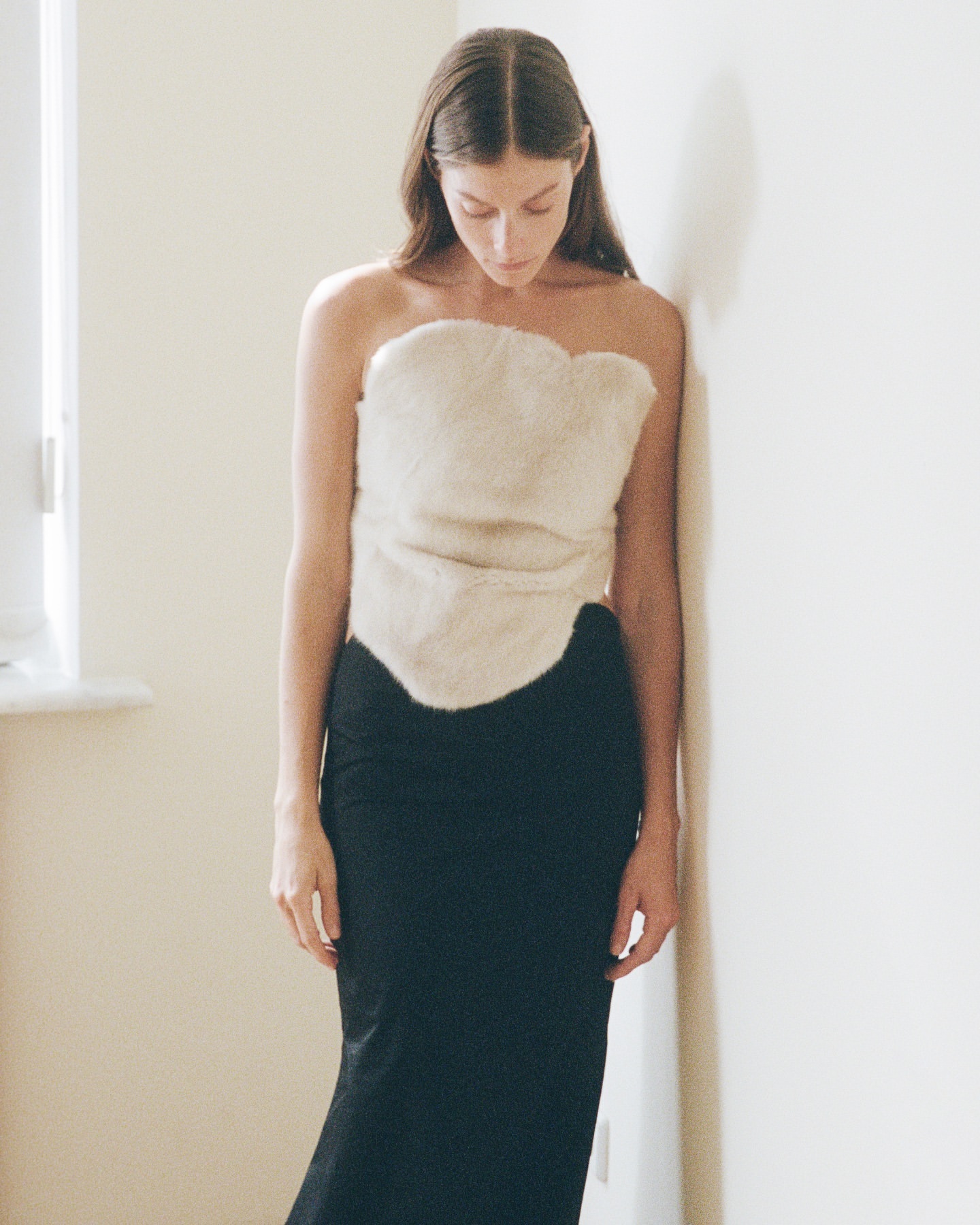 A fair-skinned woman with long brown hair poses indoors in a cream-colored faux fur top and a black maxi skirt, leaning against a white wall.