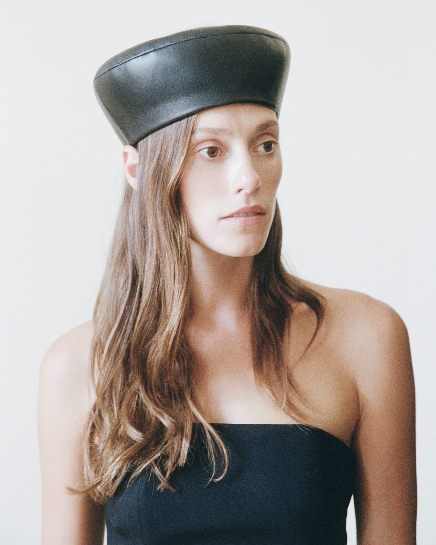 A woman with long brown hair wears a black strapless top and a black leather beret against a neutral studio background.