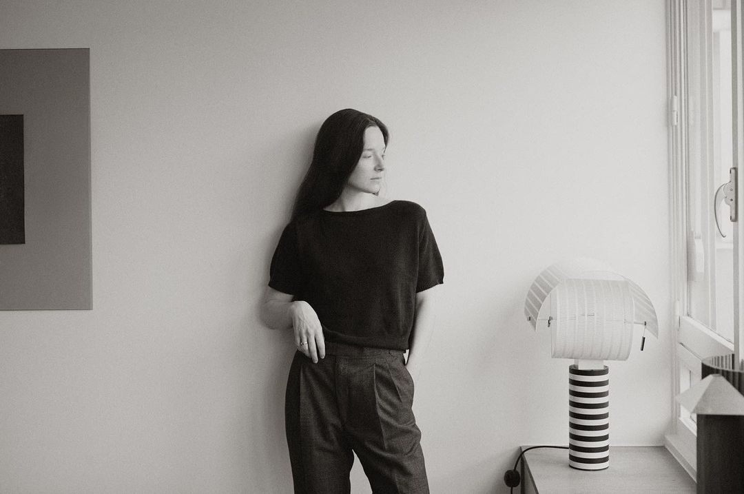 Monochrome shot of an adult woman in a dark short-sleeve shirt and high-waisted trousers leaning against a wall with a lamp and artwork.