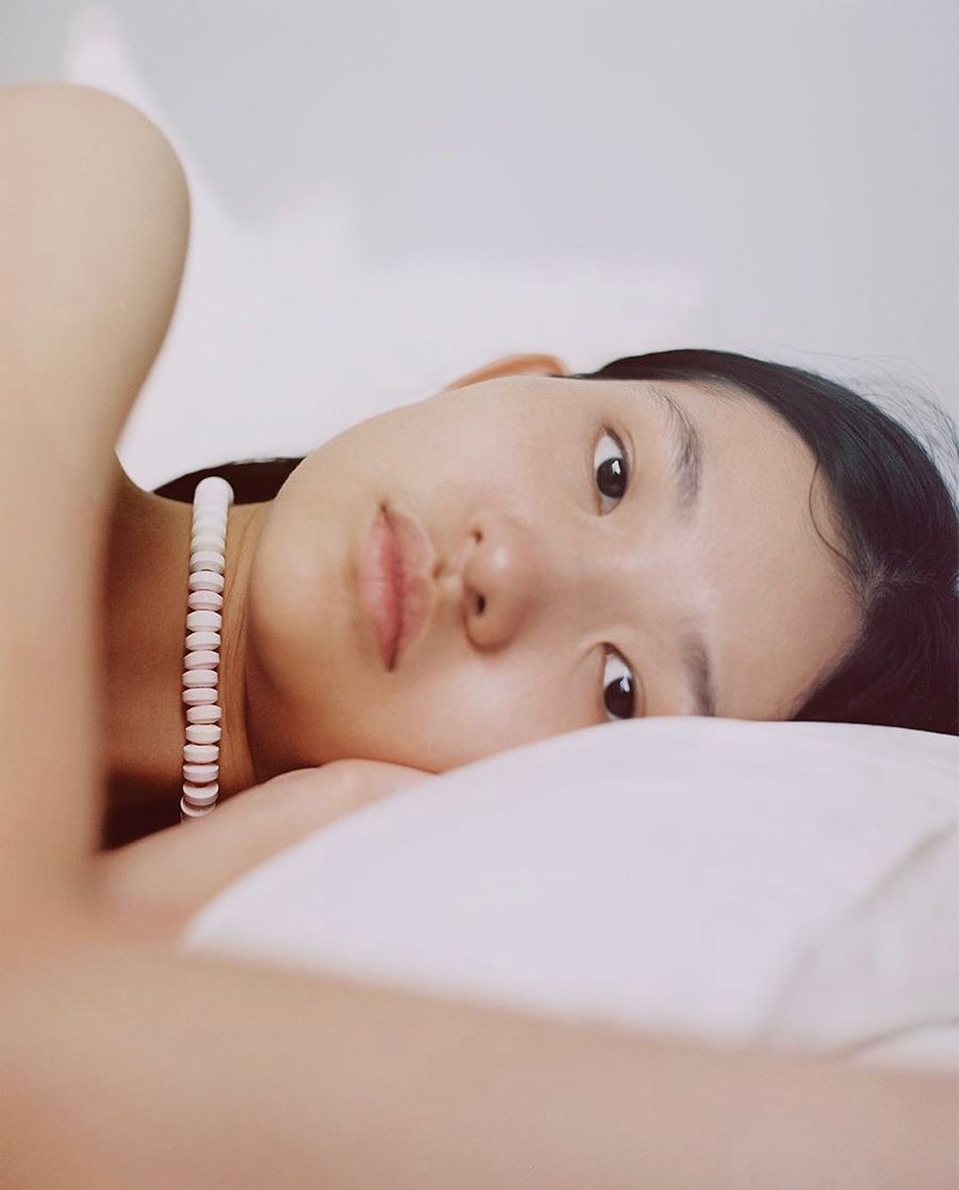 Close-up portrait of an Asian adult female lying down on a white pillow wearing a white necklace looking at the camera in an indoor setting.