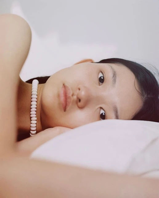 Close-up portrait of an Asian adult female lying down on a white pillow wearing a white necklace looking at the camera in an indoor setting.
