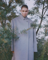 A model wearing a grey wool coat standing outdoors amidst leafy branches, photographed by Luba Kozorezova.